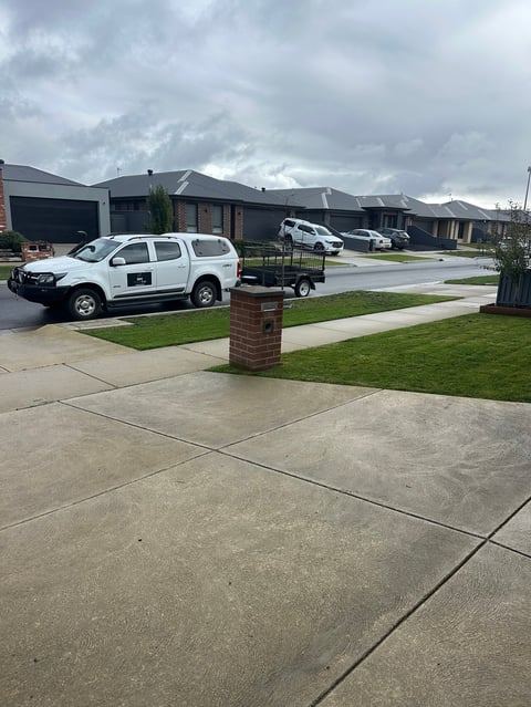 White utility vehicle parked in suburban driveway with modern houses, manicured lawn, and cloudy sky in background