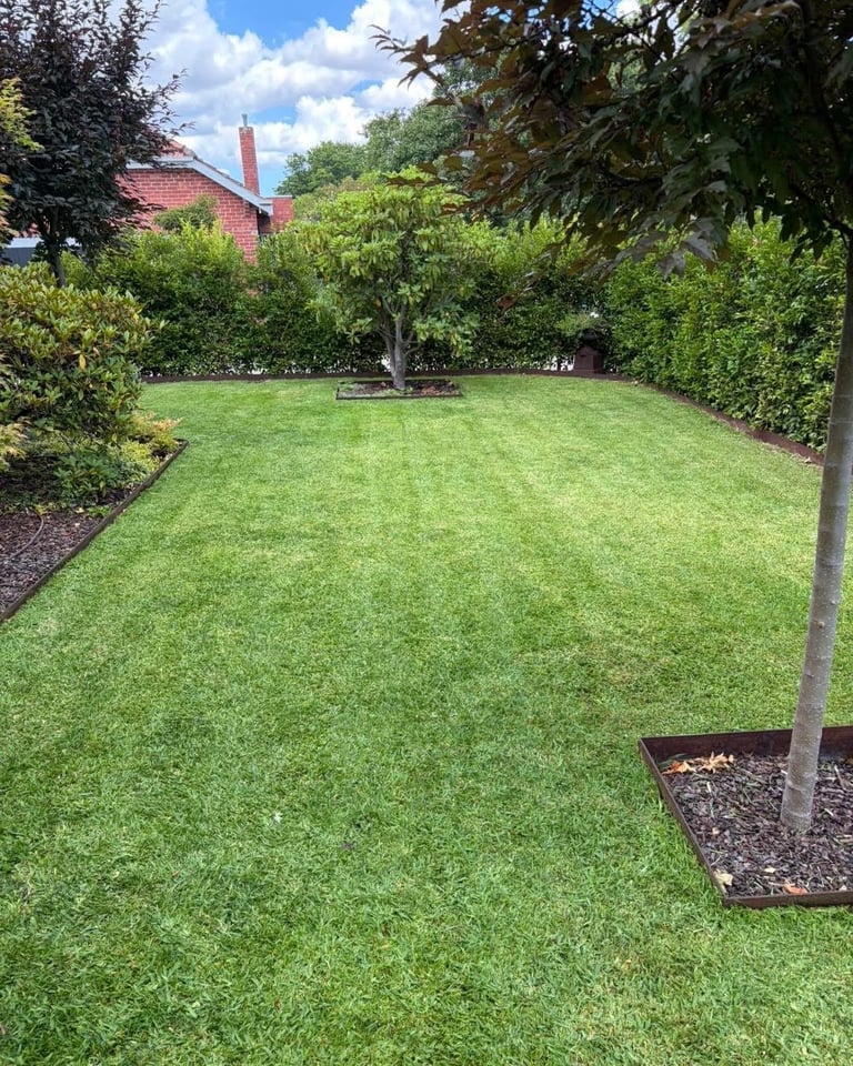 Well-maintained residential garden with lush green lawn, mature trees, hedges, and a brick house visible in the background