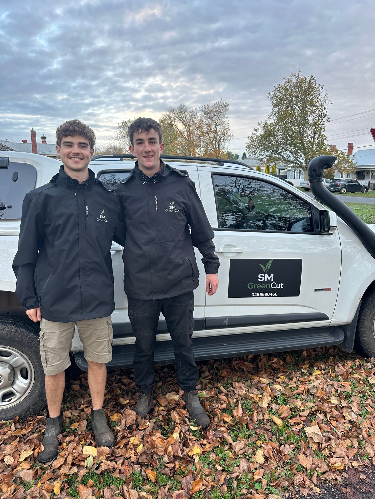 Two young workers in black jackets standing in front of a white SM GreenCut work van, surrounded by autumn leaves