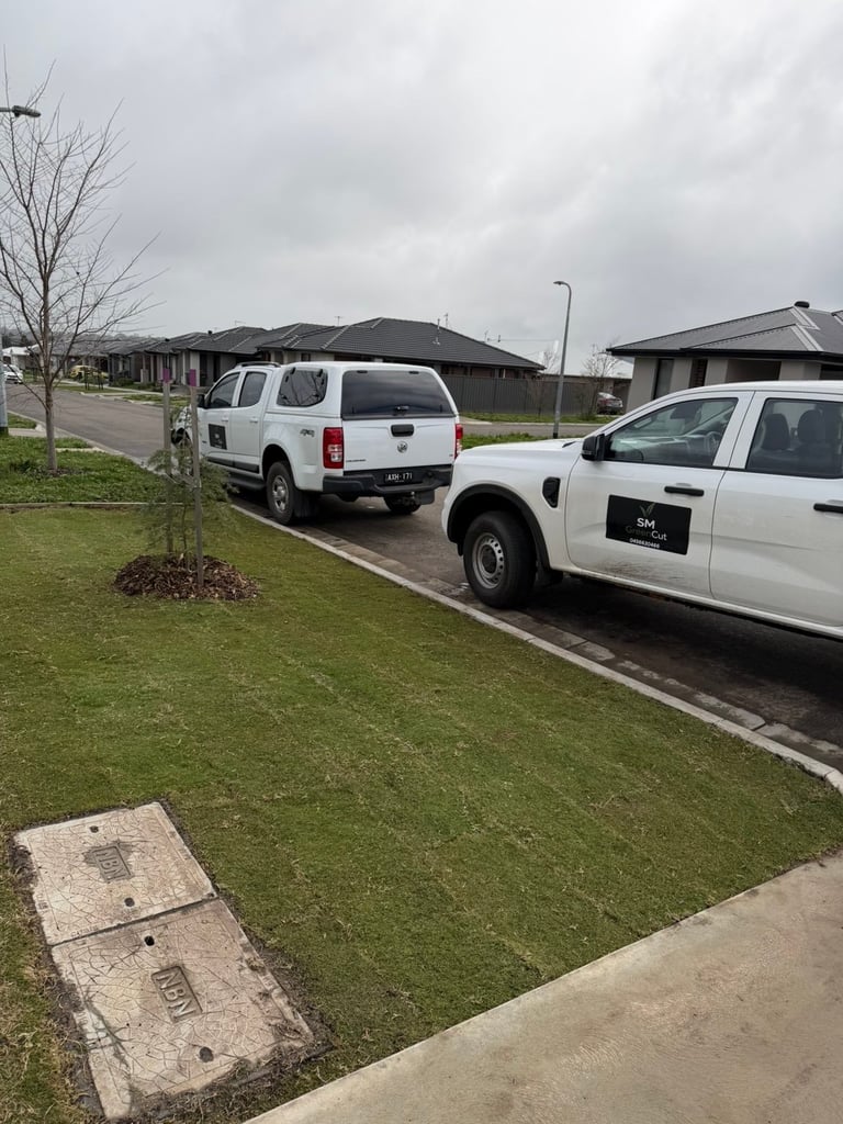 Two white utility vehicles parked on a residential street with green lawn and single-story houses in the background