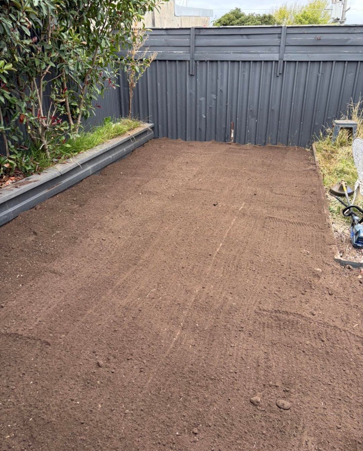 Empty backyard garden bed with brown soil, black metal fence, and raised planter box with greenery