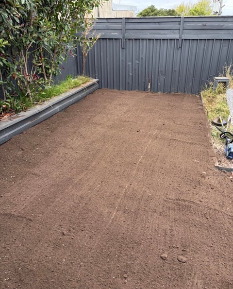 Empty residential backyard with brown soil ground, gray metal fence, and raised planter beds with vegetation