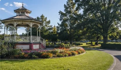 Victorian-style gazebo with red base and white railings in a sunlit park surrounded by trees and manicured lawns