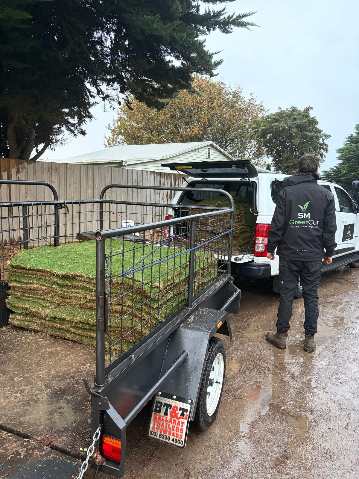 Metal livestock trailer with gate attached to white van on wet gravel driveway next to wooden fencing and trees
